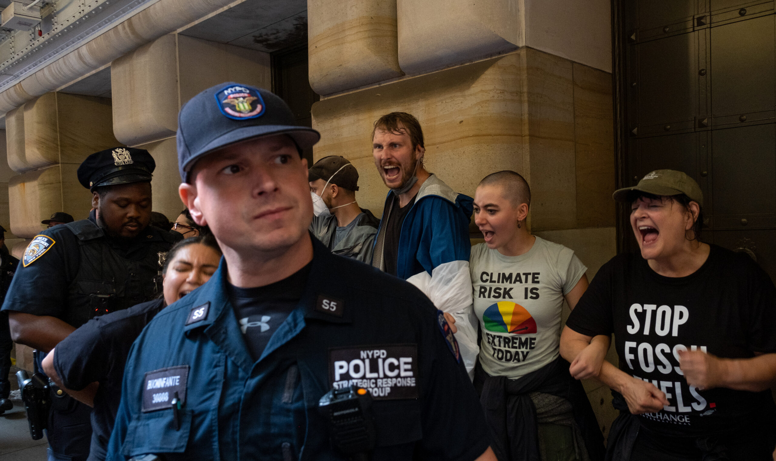 Climate protesters block the doors to the Federal Reserve Bank of New York on Monday as an NYPD police officer with the strategic Response Group, which specializes in large demonstrations, crowd control, and major events, center, watches over the demonstrators and another officer arrests a protester, left. Credit: Keerti Gopal