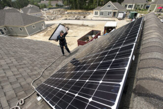 Jack Doherty, photovoltaic project manager for Revision Energy, carries a solar panel to the roof ridge of a home in OceanView at Falmouth. The company, which employs almost 200 people, has installed panels on about 50 roofs in the development. Credit: Ben McCanna/Portland Portland Press Herald via Getty Images
